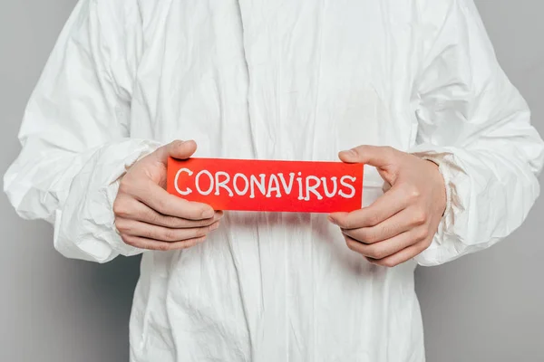 Cropped view of epidemiologist in hazmat suit holding warning card with coronavirus inscription on grey background — Stock Photo