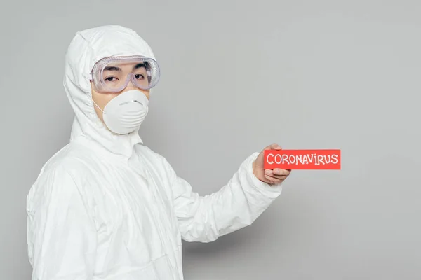 Épidémiologiste asiatique en combinaison Hazmat et masque respirateur tenant une carte d'avertissement avec inscription coronavirus sur fond gris — Photo de stock