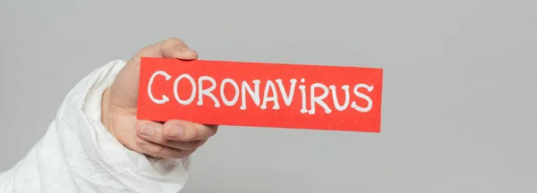 Cropped view of epidemiologist holding warning card with coronavirus inscription isolated on grey, panoramic shot — Stock Photo