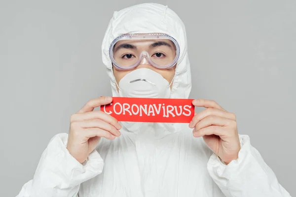 Epidemiologo asiatico in tuta hazmat e maschera respiratoria con cartellino di avvertimento con iscrizione coronavirus isolata su grigio — Foto stock