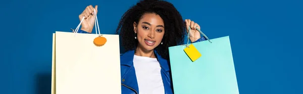 Sonriente hermosa mujer afroamericana sosteniendo bolsas de compras sobre fondo azul, tiro panorámico - foto de stock