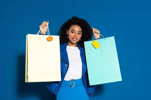 Sonriente hermosa africana americana mujer sosteniendo bolsas de compras sobre fondo azul - foto de stock