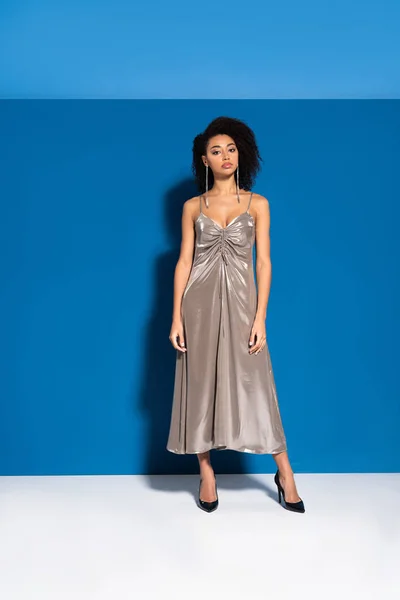 Hermosa mujer afroamericana en vestido de plata posando sobre fondo azul - foto de stock