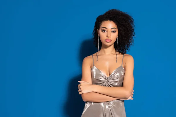 Hermosa mujer afroamericana en vestido de plata posando con los brazos cruzados sobre fondo azul - foto de stock