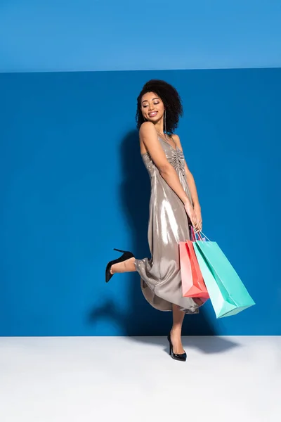 Sorrindo bela mulher americana africana em vestido de prata posando com sacos de compras no fundo azul — Fotografia de Stock