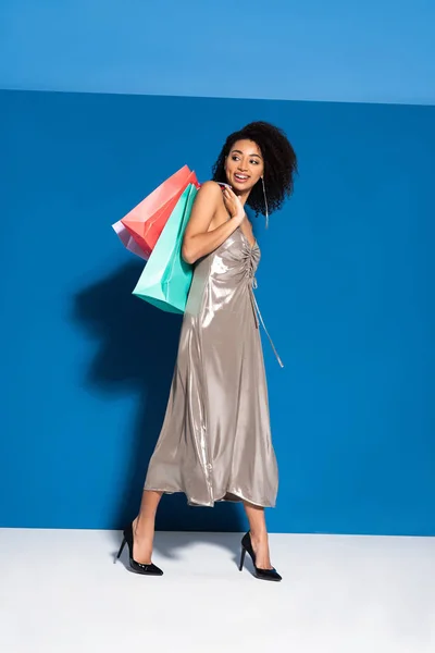 Happy beautiful african american woman in silver dress posing with shopping bags on blue background — Stock Photo