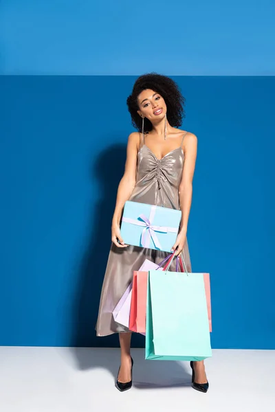 Sorrindo bela mulher afro-americana em vestido de prata segurando presente e compras em fundo azul — Fotografia de Stock
