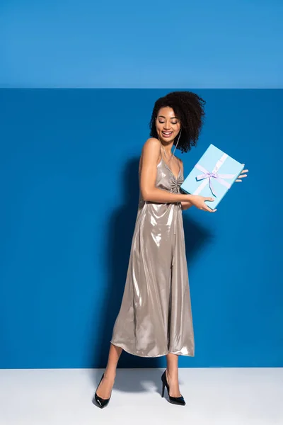 Happy beautiful african american woman in silver dress holding gift on blue background — Stock Photo