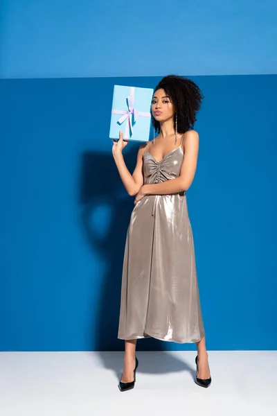 Hermosa mujer afroamericana en vestido de plata sosteniendo regalo sobre fondo azul - foto de stock