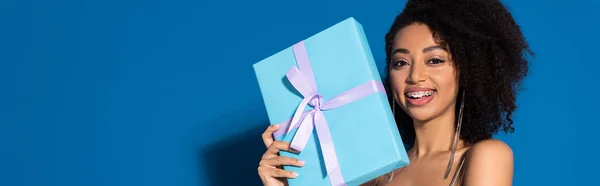 Happy beautiful african american woman holding gift on blue background, panoramic shot — Stock Photo
