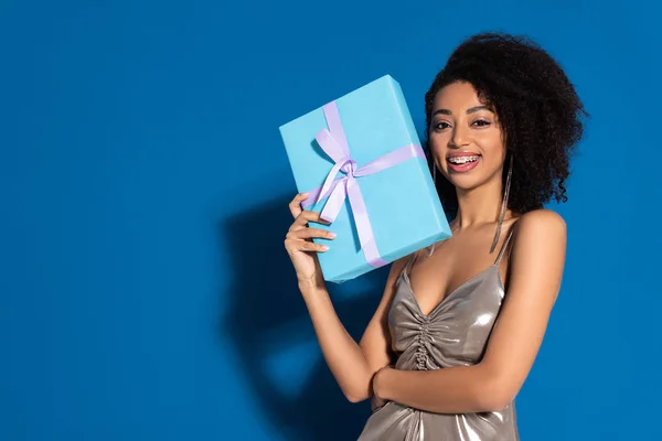 Happy beautiful african american woman in silver dress holding gift on blue background — Stock Photo