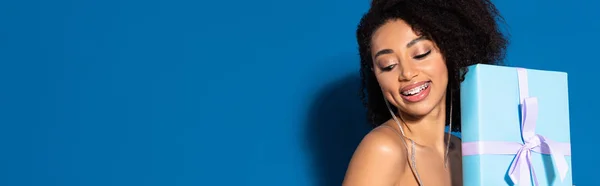 Smiling beautiful african american woman holding gift on blue background, panoramic shot — Stock Photo