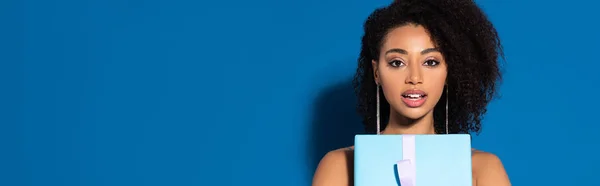 Aufgeregt schöne afrikanisch-amerikanische Frau mit offenem Mund hält Geschenk auf blauem Hintergrund, Panoramaaufnahme — Stockfoto