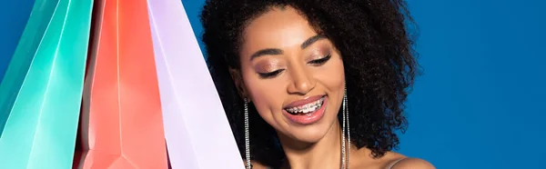 Happy beautiful african american woman holding shopping bags on blue background, panoramic shot — Stock Photo