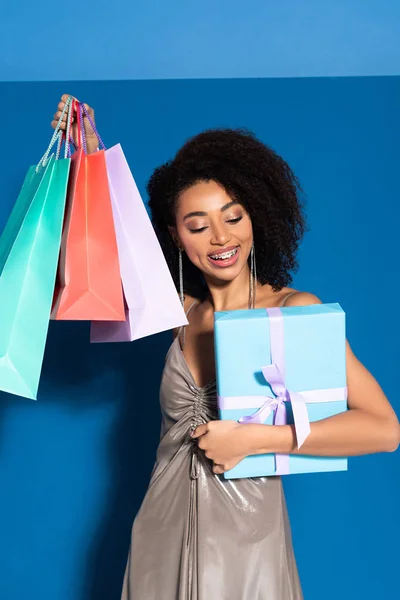 Happy beautiful african american woman in silver dress holding gift box and shopping bags on blue background — Stock Photo