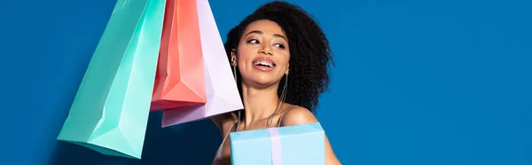 Feliz hermosa mujer afroamericana sosteniendo caja de regalo y bolsas de compras mientras mira hacia otro lado sobre fondo azul, tiro panorámico - foto de stock