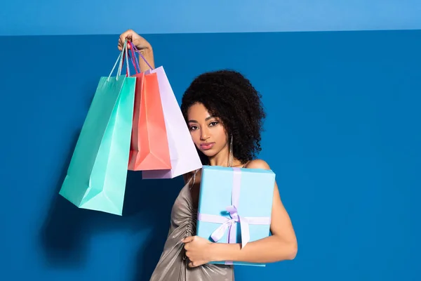 Hermosa mujer afroamericana en vestido de plata sosteniendo caja de regalo y bolsas de compras sobre fondo azul - foto de stock