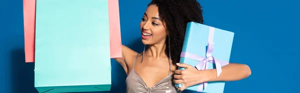 Feliz bela mulher americana africana em prata vestido segurando caixa de presente e olhando para sacos de compras em fundo azul, tiro panorâmico — Fotografia de Stock