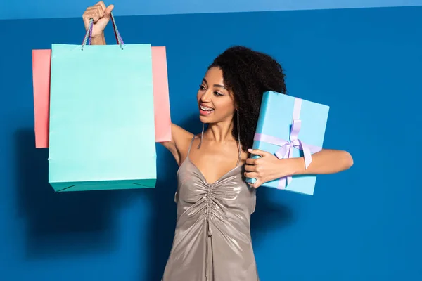 Feliz hermosa mujer afroamericana en vestido de plata sosteniendo caja de regalo y mirando las bolsas de compras sobre fondo azul - foto de stock