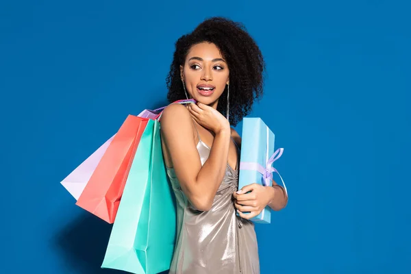 Glücklich schöne afrikanisch-amerikanische Frau in silbernem Kleid mit Geschenkschachtel und Einkaufstaschen beim Wegschauen auf blauem Hintergrund — Stockfoto