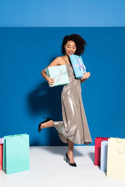 Feliz hermosa mujer afroamericana en vestido de plata sosteniendo cajas de regalo cerca de bolsas de compras sobre fondo azul — Stock Photo