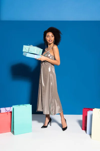 Feliz hermosa mujer afroamericana en vestido de plata sosteniendo cajas de regalo cerca de bolsas de compras sobre fondo azul — Stock Photo