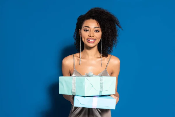Sonriente hermosa mujer afroamericana en vestido de plata sosteniendo regalos sobre fondo azul - foto de stock