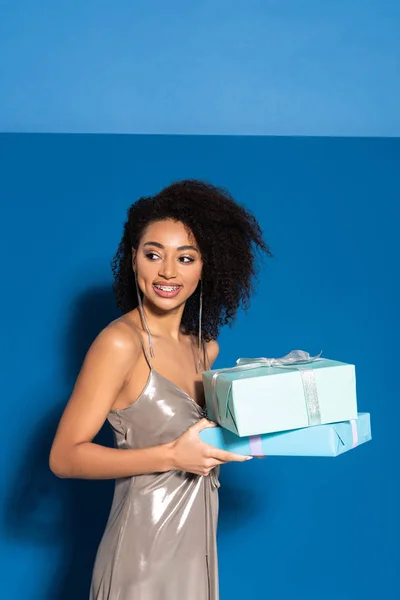 Sonriente hermosa mujer afroamericana en vestido de plata sosteniendo regalos y mirando hacia otro lado sobre fondo azul — Stock Photo