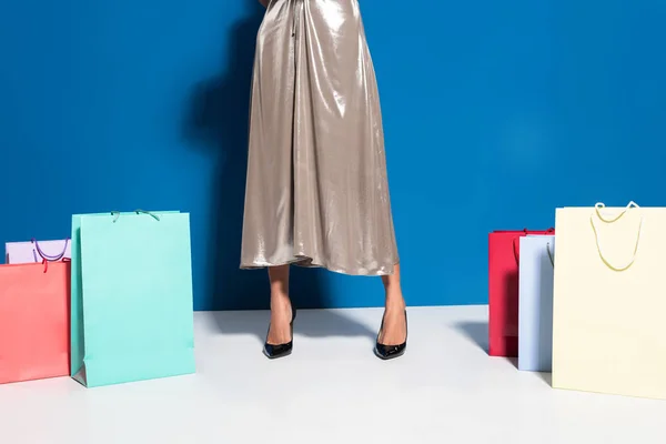 Vista recortada de mujer afroamericana en vestido de plata cerca de bolsas de compras sobre fondo azul - foto de stock