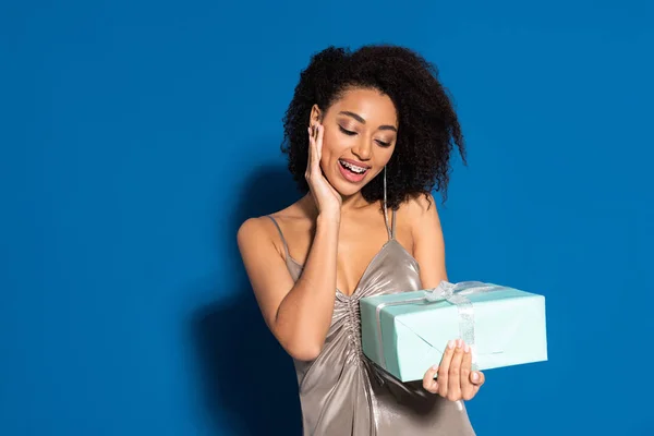 Heureuse belle femme afro-américaine en robe d'argent tenant cadeau sur fond bleu — Photo de stock