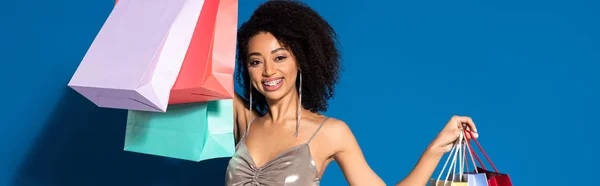 Sorrindo elegante mulher afro-americana em vestido de prata segurando sacos de compras no fundo azul, tiro panorâmico — Fotografia de Stock