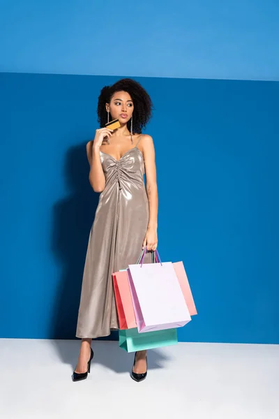 Elegant african american woman in silver dress holding shopping bags and credit card on blue background — Stock Photo