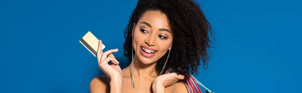 Desnudo feliz elegante afroamericana mujer sosteniendo bolsas de compras y tarjeta de crédito sobre fondo azul, tiro panorámico — Stock Photo