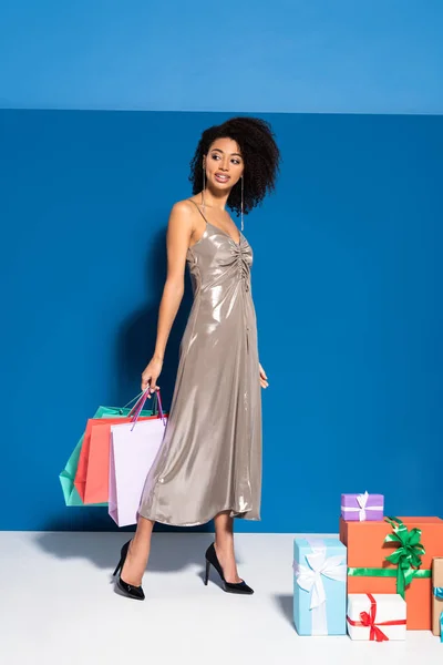 Sonriente hermosa mujer afroamericana en vestido de plata sosteniendo bolsas de compras cerca de regalos sobre fondo azul - foto de stock