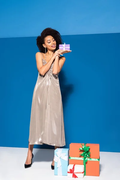 Happy beautiful african american woman in silver dress with gifts on blue background — Stock Photo