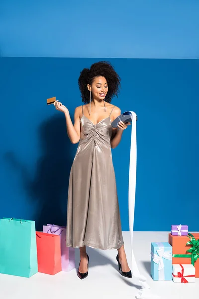 Heureuse belle femme afro-américaine en robe d'argent tenant terminal de paiement avec chèque et carte de crédit près de cadeaux et sacs à provisions sur fond bleu — Photo de stock