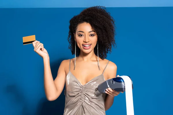 Щаслива красива афроамериканська жінка в срібній сукні тримає платіжний термінал з чековою та кредитною карткою на синьому фоні — стокове фото
