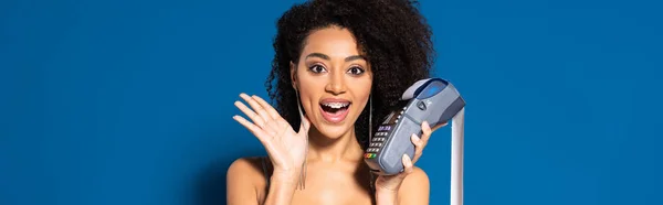 Impactado hermosa mujer afroamericana en vestido de plata que sostiene terminal con cheque sobre fondo azul, tiro panorámico - foto de stock