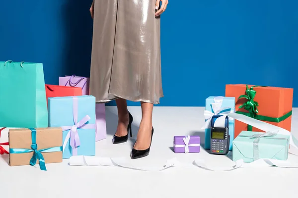 Ausgeschnittene Ansicht einer Afroamerikanerin im silbernen Kleid in der Nähe des Zahlungsterminals mit Scheck, Einkaufstaschen und Geschenken auf blauem Hintergrund — Stockfoto