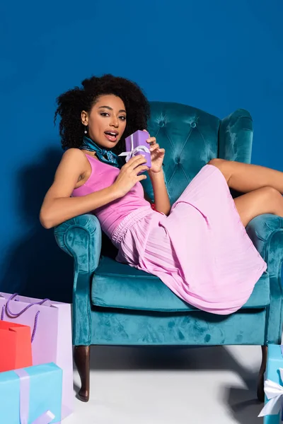 Surprised african american woman sitting in velour armchair with gift box near purchases on blue background — Stock Photo