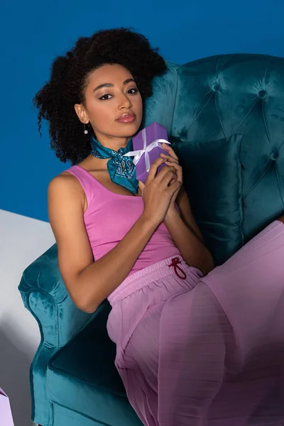 Beautiful african american woman sitting in velour armchair with gift box on blue background — Stock Photo