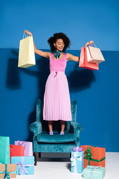 Улыбающаяся африканская американка стоящая на велюровом кресле с пакетами для покупок рядом с подарками на синем фоне — стоковое фото