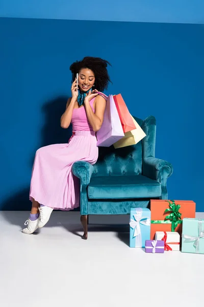 Donna afroamericana sorridente seduta in poltrona di velluto con acquisti e chiacchiere su smartphone vicino a regali su sfondo blu — Foto stock