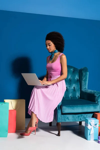 Afro-américaine assise dans un fauteuil en velours avec ordinateur portable près de boîtes-cadeaux et des achats sur fond bleu — Photo de stock