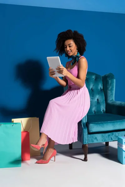 Sorrindo mulher afro-americana sentada em poltrona de veludo com tablet digital perto de compras em fundo azul — Fotografia de Stock
