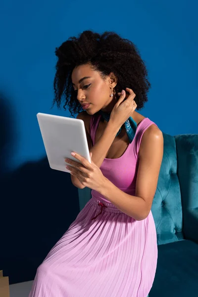 Африканская американка, сидящая в велюровом кресле с цифровой табличкой на синем фоне — стоковое фото