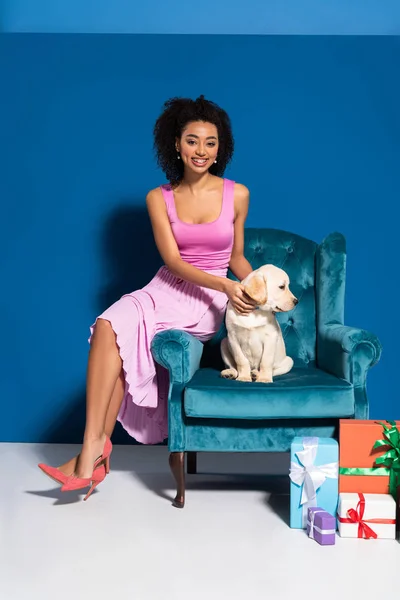 Sorrindo afro-americana mulher sentada em poltrona de veludo com filhote de cachorro golden retriever perto presentes no fundo azul — Fotografia de Stock
