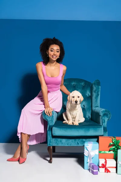 Smiling african american woman sitting in velour armchair with golden retriever puppy near gifts on blue background — Stock Photo