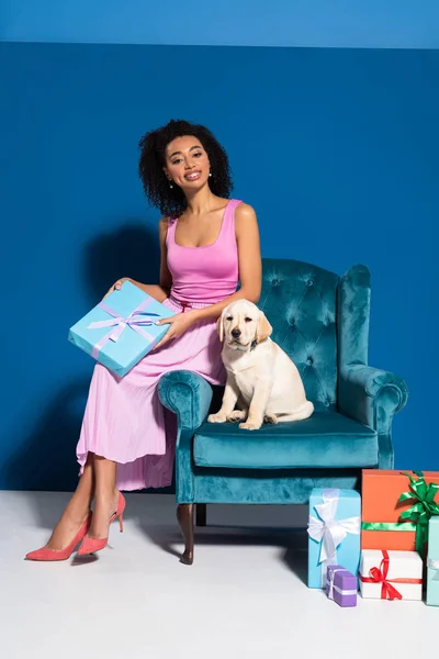 Sonriente mujer afroamericana sentada en un sillón de terciopelo con cachorro golden retriever cerca de regalos sobre fondo azul - foto de stock