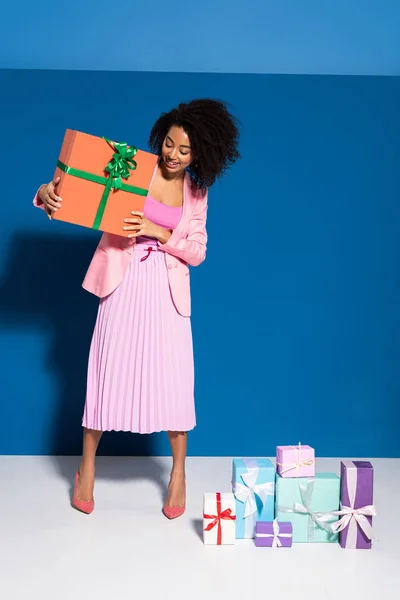 Элегантная улыбающаяся африканская американка рядом с подарками на синем фоне — стоковое фото